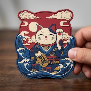 May include: A rubberized patch featuring a white cat with a blue ear, wearing a dark blue patterned robe with a gold bell. The cat holds a gold coin with black text, set against a background of dark blue ocean waves, a red sky, and clouds.
