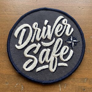 May include: A circular patch with the words "Driver Safe" embroidered in white script on a gray background. A small compass rose is to the right of the text. The patch has a navy blue border.