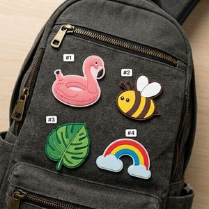 May include: A dark gray canvas backpack featuring four decorative rubber charms attached to the front pocket. The charms include a pink flamingo pool float labeled #1, a yellow and black bee labeled #2, a green monstera leaf labeled #3, and a rainbow with clouds labeled #4.