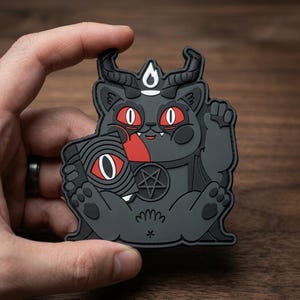 May include: A dark gray patch featuring a cartoon cat with horns, red eyes, and a pentagram. The cat is holding a book with an eye symbol and has a flame above its head. The patch is held by a hand.