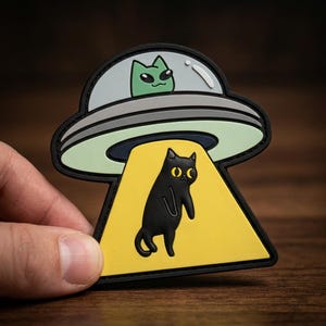 May include: A black cat is being abducted by a UFO patch. The UFO is gray and white with a green alien inside. The cat is standing in a yellow beam of light. The patch has a black border.