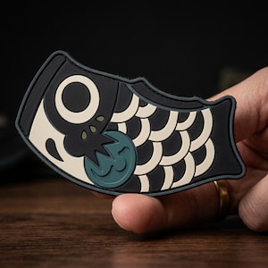 May include: A rubberized patch shaped like a stylized fish, featuring a black and cream-colored scale pattern, a large circular eye, and a teal-colored circular accent near the head, designed with a flat, matte finish and raised edges.