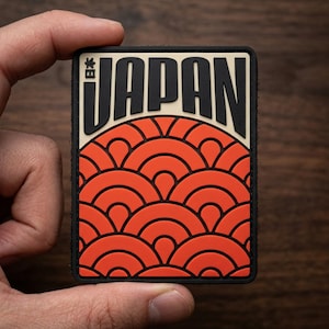 May include: A rectangular PVC patch featuring the word JAPAN in bold black lettering above a stylized orange and black wave pattern. The design includes the Japanese kanji for Japan in the top left corner, all set against a cream background.