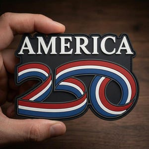 May include: A black patch with the word "AMERICA" in white block letters above the number "250" in red, white, and blue ribbon design. The patch is held in a hand against a wooden background.