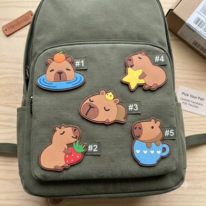 May include: An olive green canvas backpack featuring five distinct capybara PVC patches labeled #1 through #5. The patches depict capybaras with an orange, a strawberry, a yellow star, a small bird, and a blue mug with white hearts.