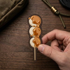 May include: A sticker shaped like a skewer of three round dango dumplings. Each off-white dumpling is topped with a glossy, brown, semi-translucent glaze. The sticker features a dark brown outline and a thin, light-colored stick at the bottom.