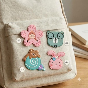 May include: A beige canvas backpack featuring four decorative patches: a pink flower with a girl's face, a green bear-like character with glasses, a brown bear sitting on a teal swirl lollipop, and a pink rabbit with a blue flower on its head.