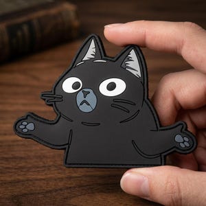 May include: A black cat-shaped patch with a surprised expression. The cat has large white eyes, gray accents on the ears, and outstretched paws. The patch is held by a hand against a blurred wooden background.