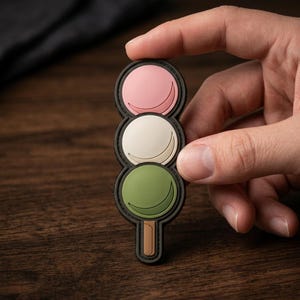 May include: A three-tiered dango-shaped patch featuring a pink, white, and green circular design stacked vertically. Each circle contains a crescent-shaped indentation, and a small brown rectangular base extends from the bottom of the green circle.