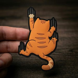 May include: Orange cat-shaped sticker with brown stripes and a black outline. The cat is depicted scratching with its claws, with claw marks on a black background. The sticker is held by a hand.
