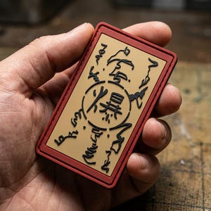 May include: A rectangular patch with a tan center and a dark red border. The center features a black circular emblem containing the Japanese kanji for explosion, surrounded by additional black stylized markings and symbols across the tan surface.