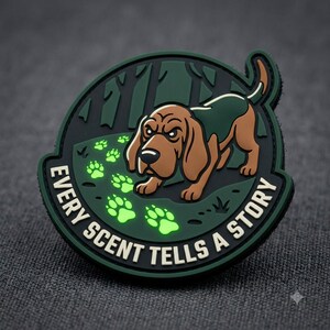 May include: A circular morale patch featuring a brown and green hound dog tracking glowing light green paw prints in a dark forest. The text EVERY SCENT TELLS A STORY is printed in white capital letters along the bottom edge of the dark green border.