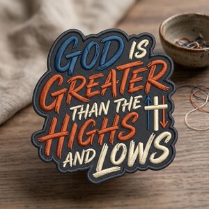 May include: Embroidered patch with the text "GOD IS GREATER THAN THE HIGHS AND LOWS" in blue, orange, and white on a dark gray background. The patch has a decorative border and is likely for clothing or accessories.