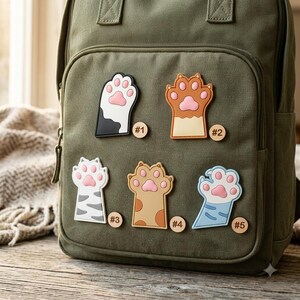May include: An olive green canvas backpack featuring five distinct cat paw patches attached to the front pocket. Each patch displays a unique color pattern and is labeled with a small circular wooden tag numbered from one to five.