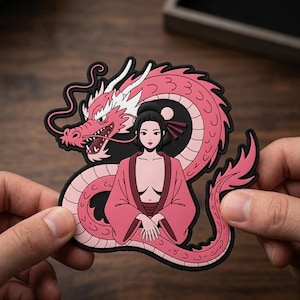May include: A flexible rubber patch featuring a pink dragon with white accents coiled behind a figure with dark hair styled in a bun, wearing a pink robe. The design is outlined in black, creating a bold graphic illustration with a smooth finish.