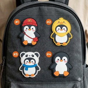 May include: Four decorative rubber patches featuring cartoon penguins attached to a dark gray backpack. Patch #1 shows a penguin in a red beanie with a red bag. Patch #2 shows a penguin in a yellow duck hood. Patch #3 shows a penguin in a panda hood with a blue scarf. Patch #4 shows a plain penguin.