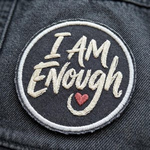 May include: Embroidered patch with the words "I AM ENOUGH" in cream-colored script, a small red heart, and a white border on a dark gray background. The patch is circular and appears to be sewn onto a denim jacket.