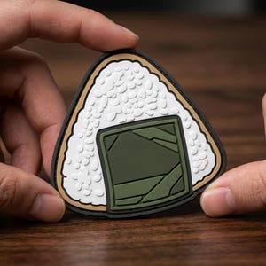 May include: A triangular-shaped patch designed to resemble an onigiri rice ball. The patch features a textured white surface representing rice grains, accented by a dark olive green square section with geometric lines, all bordered in black.