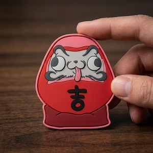 May include: A red and pink Daruma-style patch featuring a cartoon cat face with wide eyes, a protruding tongue, and dark gray eyebrows. The center of the patch displays the black Japanese kanji character for good fortune, known as Kichi.