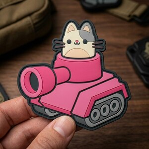 May include: A pink tank-shaped patch with a cartoon cat peeking out of the turret. The cat has a beige face, gray ear, and black whiskers. The patch has a black border and is on a wooden surface.