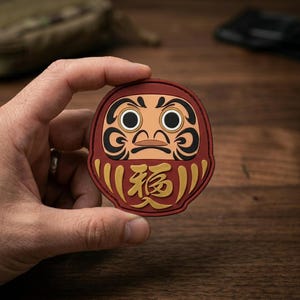 May include: A circular patch featuring a stylized Daruma doll design in red, black, and beige. The doll has large white eyes with black pupils, black eyebrows, and a gold-colored Japanese kanji character for luck centered on its lower body.
