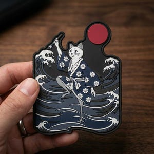 May include: A rectangular rubber patch featuring a white cat wearing a dark blue kimono with white flower patterns. The cat holds a brush against a background of stylized dark blue ocean waves and a solid red circle representing the sun.