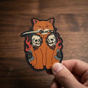 May include: A rubber patch featuring an orange cat with a scar over one eye, holding a katana in its mouth. The cat is surrounded by red flames, grey cloud patterns, and two beige human skulls. The design has a black border and flat finish.