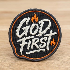 May include: Embroidered patch with the words "GOD FIRST" in white script on a black background. The patch has an orange border and small orange flame graphics. The patch is circular in shape.