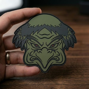 May include: A rubberized morale patch featuring a stylized, olive green creature face with a sharp, beak-like nose, dark hair, and narrow eyes with red pupils. The design is outlined in black, creating a bold, graphic appearance for this accessory.