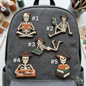 May include: Five skeleton-themed patches attached to a gray canvas backpack. Each skeleton is depicted in a different pose with books, labeled #1 through #5. The designs feature skeletons wearing hats or flowers, reading, or holding books with the text READ and BOOKS.