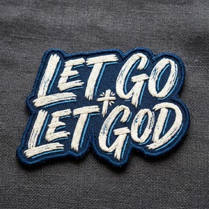 May include: Embroidered patch with the words "LET GO LET GOD" in white script on a navy blue background. A small cross is between the words. The patch has a navy blue border and is on a gray fabric.