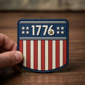 May include: A shield-shaped patch with a patriotic design. The top section is navy blue with white stars and the year "1776". Below are red and white vertical stripes. The patch is held by a hand.