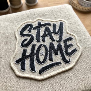 May include: Embroidered patch with the words "STAY HOME" in black thread on a beige fabric background. The patch has a cream-colored border and a small star detail. The patch is likely for sewing onto clothing or other fabric items.