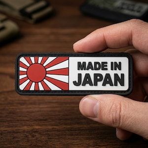 May include: A rectangular patch with a black border featuring a red and white rising sun graphic on the left side and the text MADE IN JAPAN in bold black lettering on a white background on the right side of the rectangular patch.