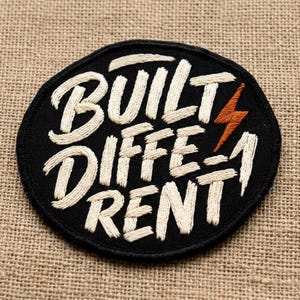 May include: A black, oval-shaped embroidered patch with the words "BUILT DIFFERENT" in cream-colored thread. An orange lightning bolt is to the right of the word "DIFFERENT". The patch is on a tan, textured background.