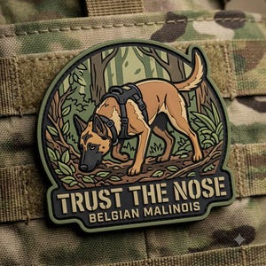 May include: A rubberized morale patch featuring a tan Belgian Malinois dog wearing a black harness, depicted in a forest setting. The text "TRUST THE NOSE" and "BELGIAN MALINOIS" is printed in a bold, stencil-style font at the bottom of the patch.