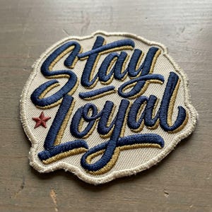 May include: Embroidered patch with the words "Stay Loyal" in navy blue script with gold accents. A small red star is on the left side. The patch has a beige background and a stitched border.