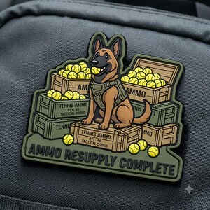 May include: A tactical morale patch featuring a brown German Shepherd wearing a harness, sitting on wooden crates filled with yellow tennis balls. The crates are labeled TENNIS AMMO QTY: 48 TACTICAL DOGGO. The text AMMO RESUPPLY COMPLETE is at the bottom.