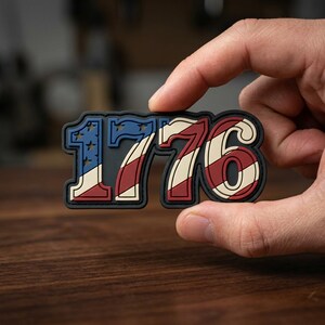 May include: A black patch with the number "1776" in red, white, and blue, resembling the American flag. The "1" and "7" are red and white striped, while the top of the first "7" has a blue background with white stars.