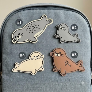 May include: Four seal-shaped patches are arranged on a gray fabric surface. Patch #1 is a large gray spotted seal. Patch #2 is a smaller gray seal. Patch #3 is a brown seal with a dark line detail. Patch #4 is a cream-colored seal.