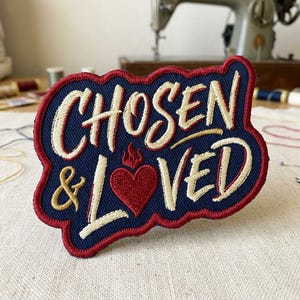 May include: Embroidered patch with the words "CHOSEN & LOVED" in cream-colored script on a navy blue background. A red heart with flames is centered below the word "CHOSEN". The patch has a red border.