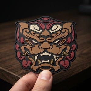 May include: A molded rubber patch featuring a stylized, symmetrical tiger face design in black, brown, cream, and dark red. The intricate graphic depicts a fierce feline expression with sharp teeth, pointed ears, and swirling patterns throughout.