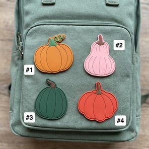 May include: A sage green canvas backpack featuring four pumpkin-shaped patches labeled #1, #2, #3, and #4. Patch #1 is orange, #2 is light pink, #3 is dark green, and #4 is red-orange. Each patch has a brown stem and distinct vertical ridges.