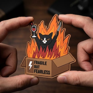 May include: A patch featuring a black cat with a white chest and face, holding a wine glass and a knife, surrounded by flames. The cat is in a cardboard box with the text "FRAGILE BUT FEARLESS" and a lightning bolt.