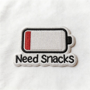 May include: Embroidered patch featuring a white battery outline with a red bar indicating low power. The patch has a black border and the words "Need Snacks" in black script below the battery symbol.