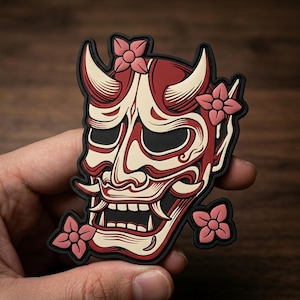 May include: A rubberized patch featuring a stylized Hannya mask in cream, red, and black. The mask has prominent horns, sharp teeth, and deep eye sockets, accented by four pink four-petaled flowers positioned around the perimeter of the design.
