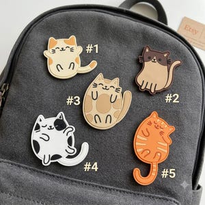 May include: Five cat-shaped rubber patches attached to a dark gray canvas backpack. The patches include a cream and orange cat labeled #1, a brown and tan cat labeled #2, a tan spotted cat labeled #3, a white and black cat labeled #4, and an orange striped cat labeled #5.