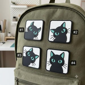 May include: An olive green canvas backpack featuring four square patches with black cat illustrations. Each patch displays a black cat with teal eyes on a white background, labeled with numbers #1, #2, #3, and #4 in black text next to each.