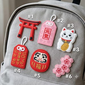 May include: A collection of six rubberized patches featuring a red torii gate, a pink omamori charm with Japanese text, a white beckoning cat, a red omamori charm with Japanese text, a red Daruma doll, and a cluster of pink cherry blossom flowers.