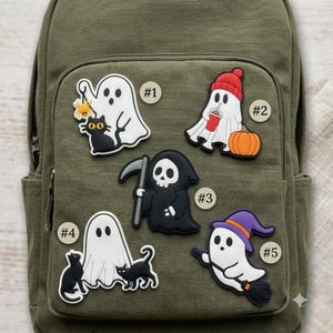 May include: Five Halloween-themed rubber patches attached to an olive green backpack. The designs include a ghost with a spider and black cat, a ghost in a red beanie with a drink and pumpkin, a grim reaper, a ghost with two black cats, and a ghost witch.
